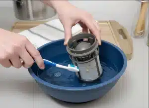 Hands cleaning a dishwasher filter with a toothbrush over a blue bowl.